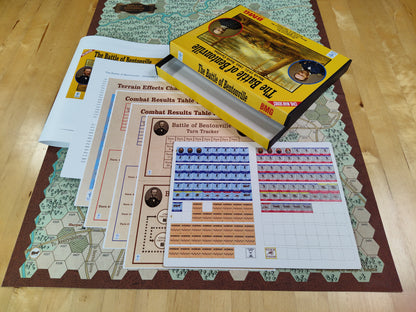 Wargaming tabletop game components including a box, maps, and charts on a wooden surface.